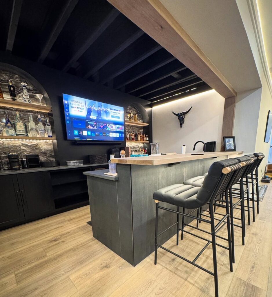 Modern home bar with dark grey counter, leather stools, and liquor shelves.
