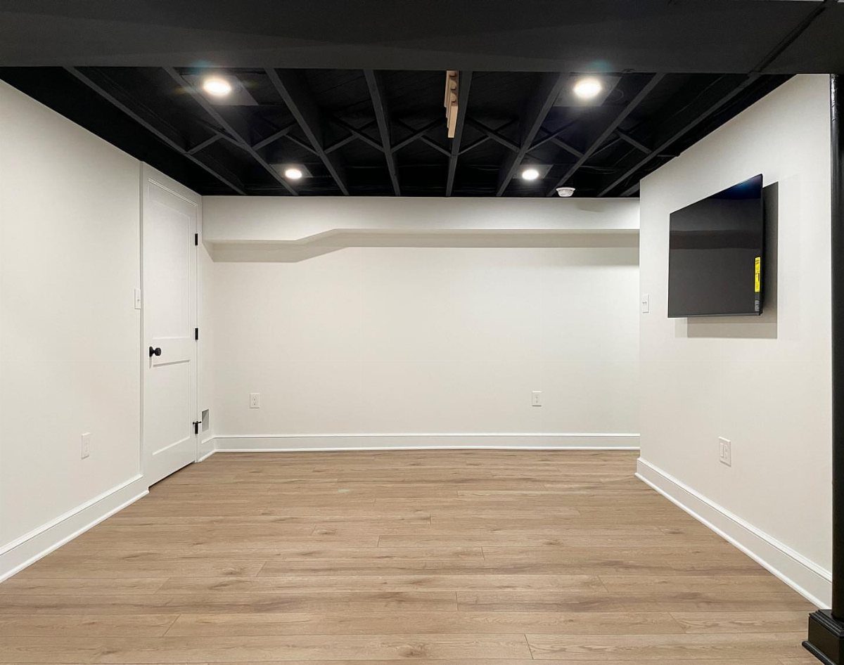 Empty basement room with white walls and an open black painted ceiling.