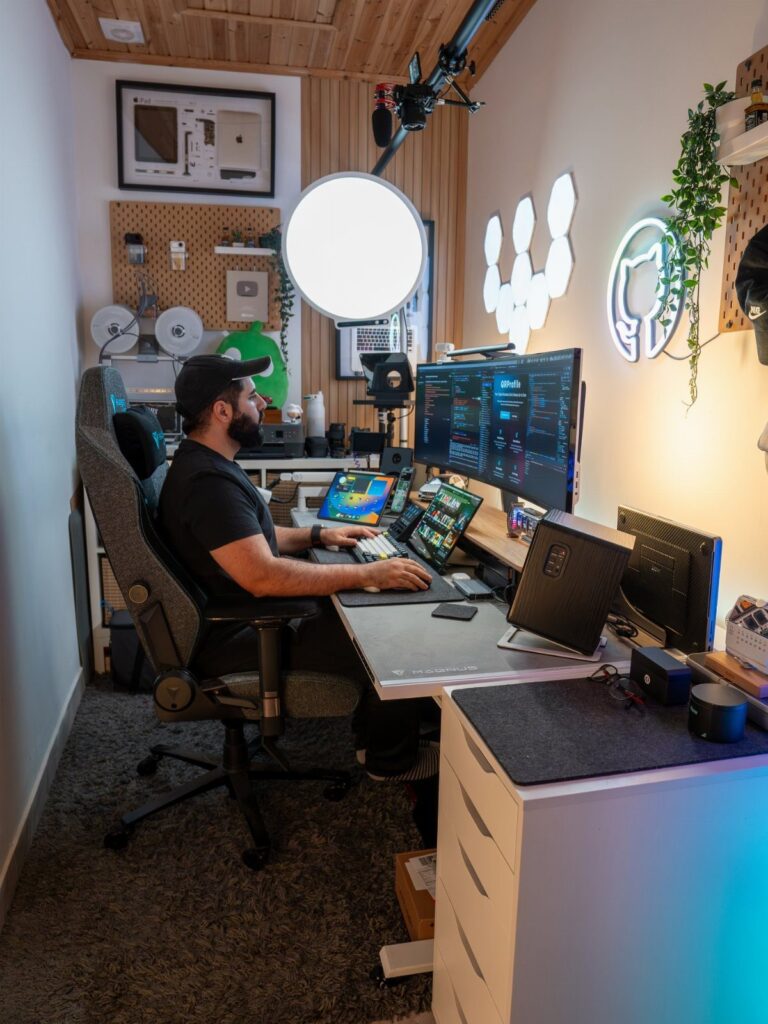 Man working at a white corner desk with multiple monitors and a ring light.