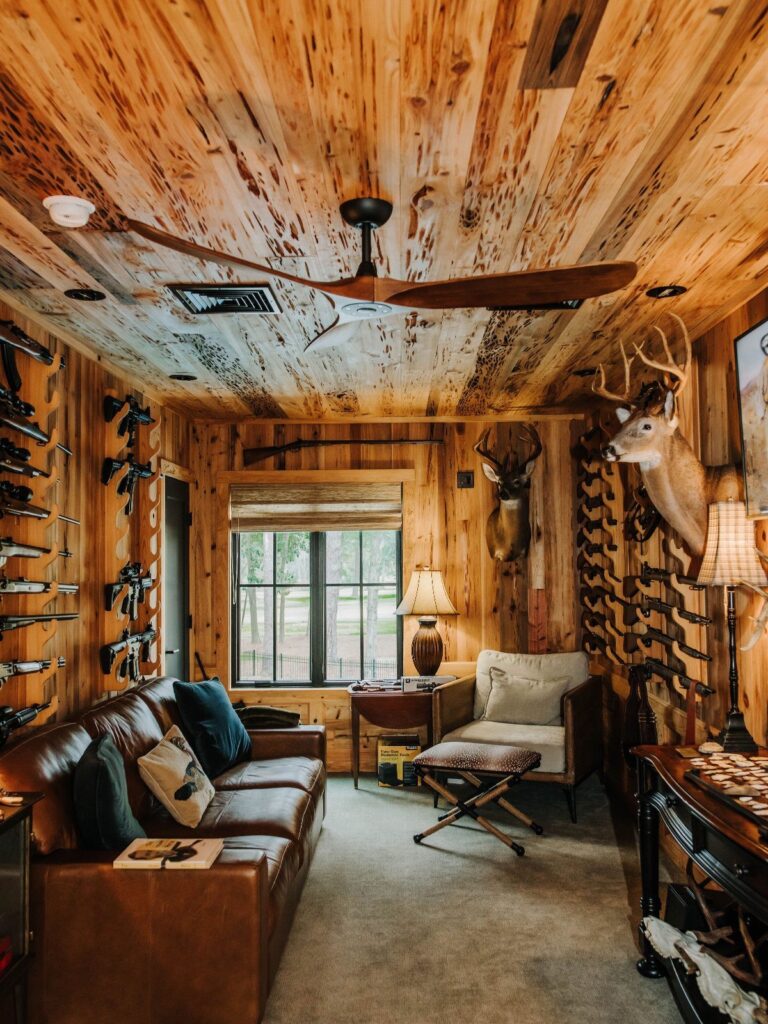 Rustic hunting room with wood paneling, leather sofa, mounted deer, and rifle rack.