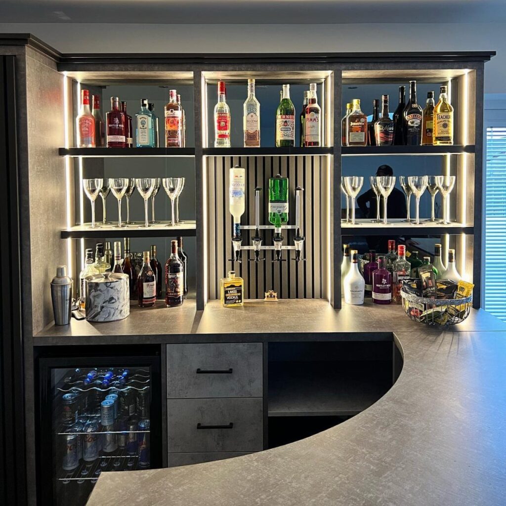 Modern home bar with grey concrete counters, backlit shelves, and a small beverage cooler.